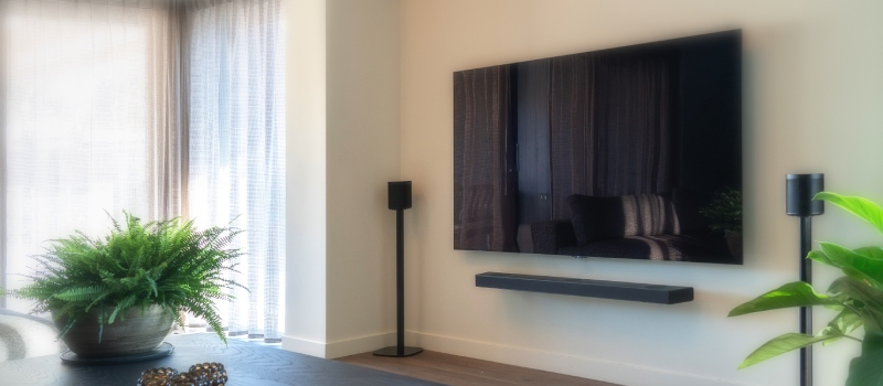 Vardagsrum med vita gardiner till vänster och en TV med soundbar och två separata högtalare på högtalarstativ på väggen till höger